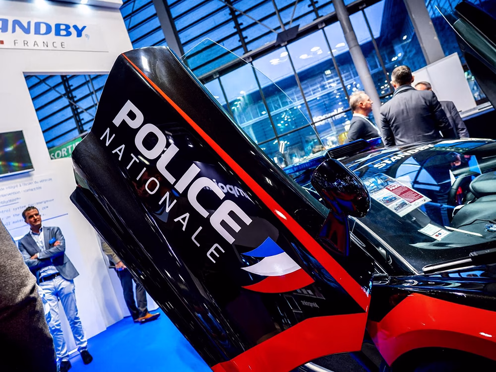 Closely cropped view of black McLaren, showing an open upward-opening door with 'Police Nationale' livery on. Mid-focus shows small groups of people in conversation on the Standby France / Standby Group stand at Milipol Paris 2025.