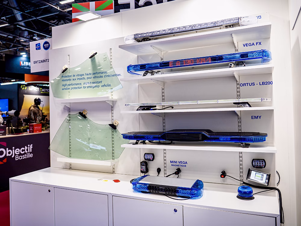 Range of unlit lightbars on white shelves and 2 pieces of glass from car windows (one broken, one unbroken) showing protective glass film on the Standby France / Standby Group stand at Milipol Paris 2025.