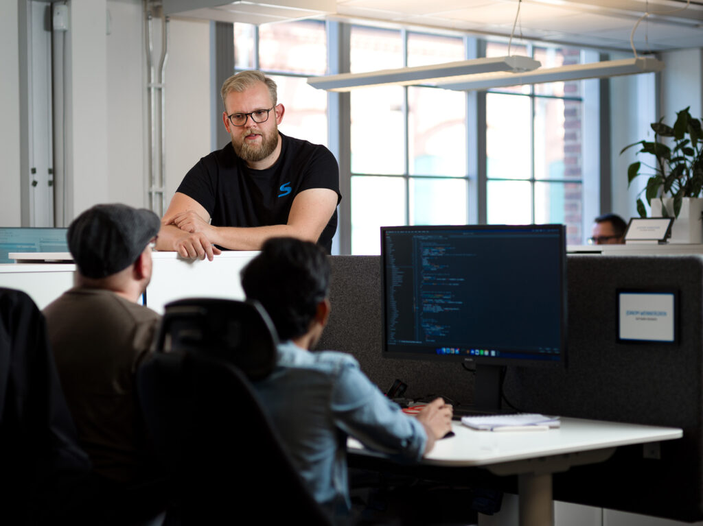 En medarbetare står vid en skärmvägg och samtalar med två andra medarbetare som sitter framför en dator.