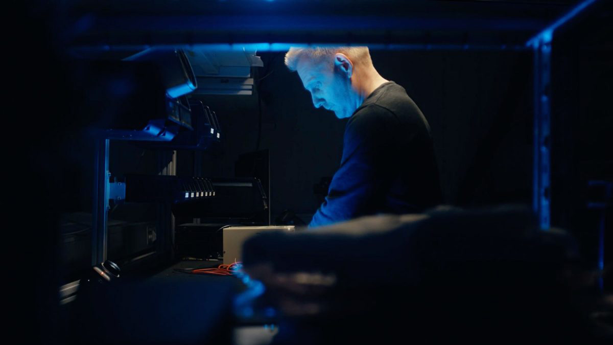 Vy från sidan på en medarbetare som står och jobbar vi en arbetsbänk i mörker med blått ljus som lyser upp.