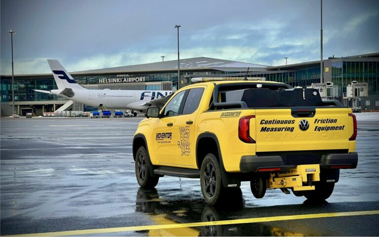 En gul pickup parkerad på en flygplats med flygplan och flygplatsbyggnad i bakgrunden.
