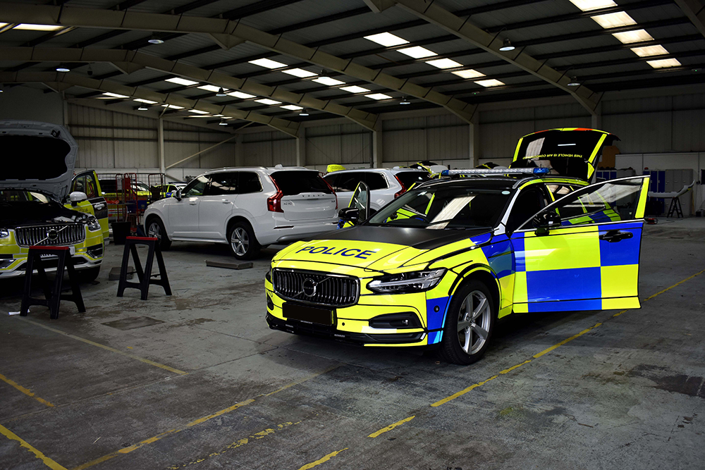 Police car at an angle view in a dark garage with doors and boots open and white cars at various stages of work in the background