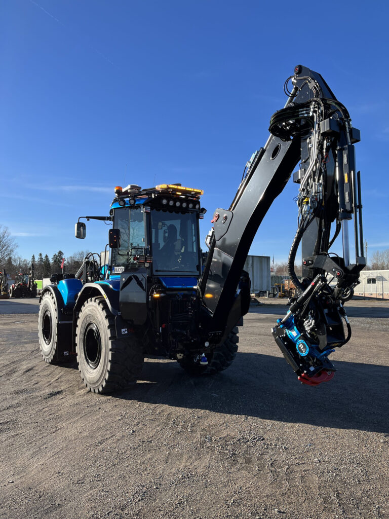 Portrait Format Angle View of Blue Excavator with Various Amber LED Modules and Amber W3 Lightbar with Blue Sky and Grey Industrial Building in Background