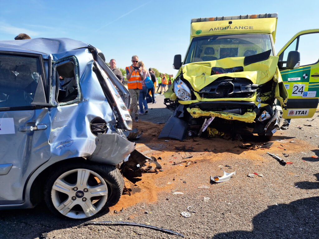 Front on shot of an ambulance involved in a controlled collision, left side door is open, bonnet smashed and left headlight missing, back end of car in lefthand foreground, boot smashed and tyre off wheel, people taking photos between the two vehicles with debris scattered on floor