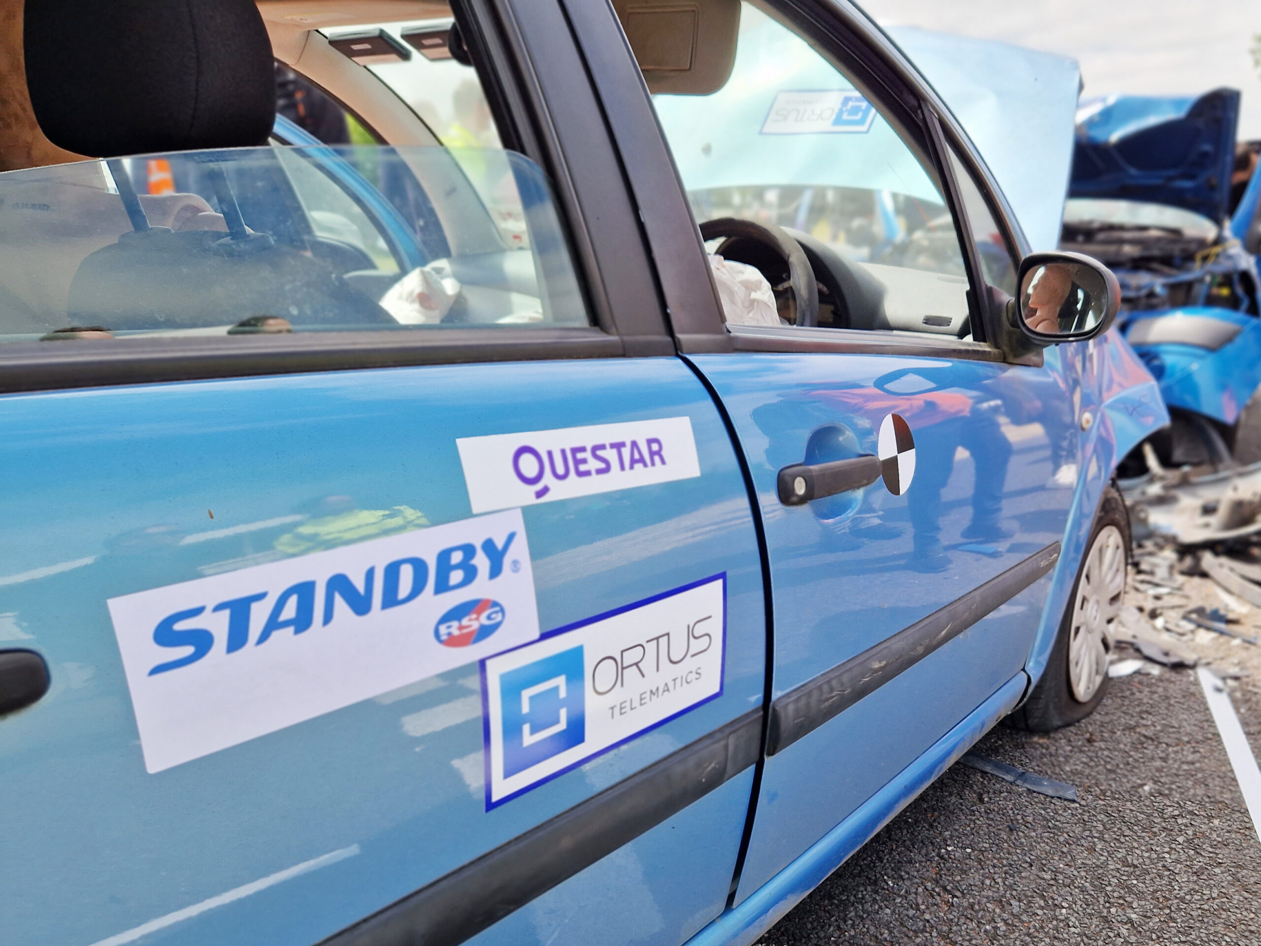 Close up of light blue car with Standby UK logo on white signage on side, the view is at an angle, the bonnet is up, another darker blue car is out of focus in the background showing signs of impact and debris.