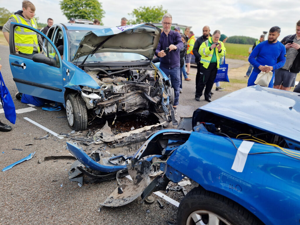 Angle view of 2 blue cars, one light and one darker, that have been subjected to a controlled head on collison. The bumper is completely detached from the light blue vehicle with multiple signs of impact on the darker one, debris is scattered around and people are crowded around observing and taking photos.