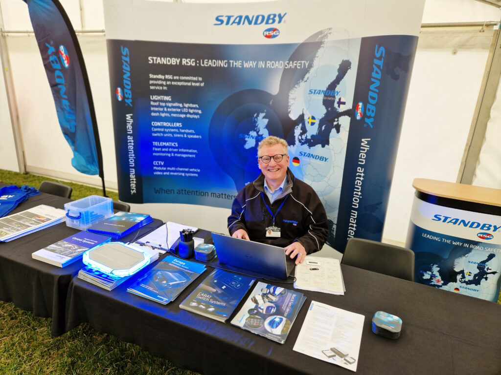 Manager wearing Standby UK uniform is sitting at table in front of Standby UK signage and smiling at camera, looking up from laptop. Various brochures and Standby UK branded products are pilled on the table.