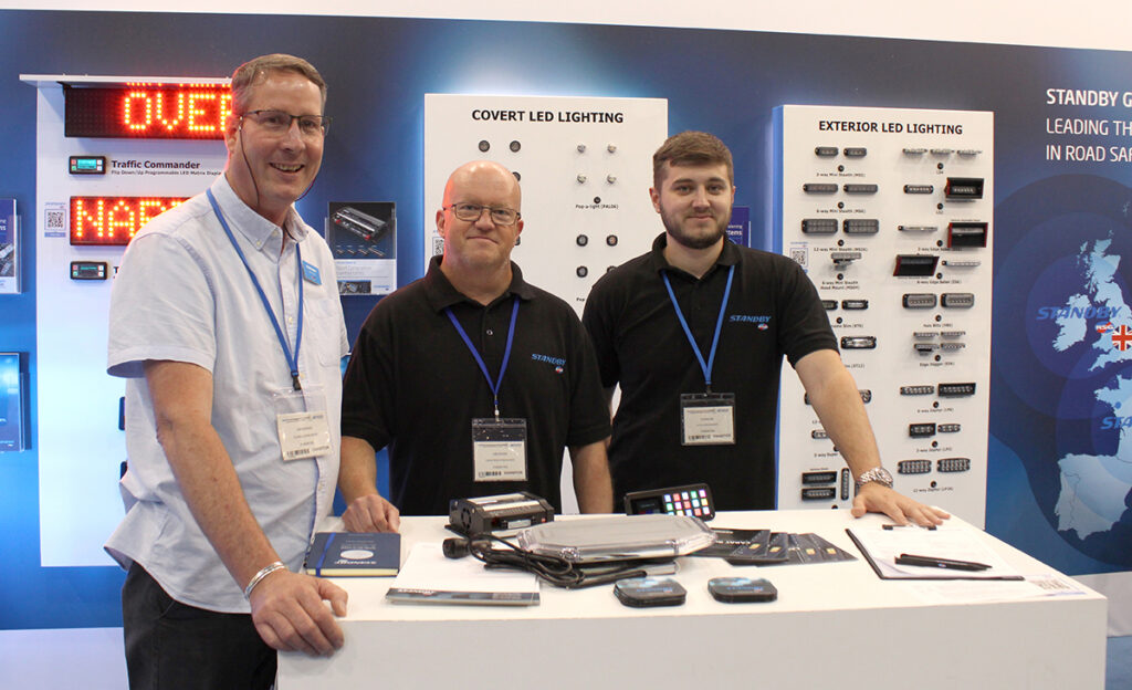 Front view of staff on Standby's NAPFM stand, three workers stand behind centre table, leaning on it with hands, looking and smiling at the camera