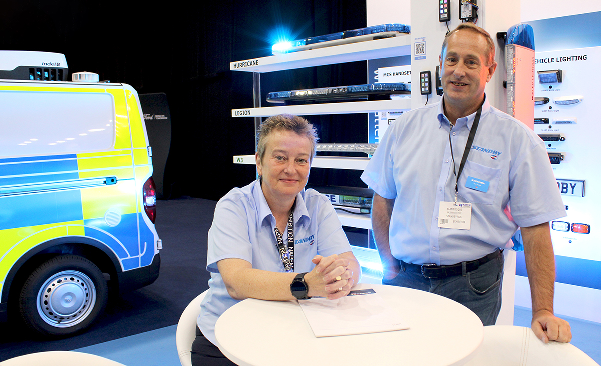 Staff at Standby stand at NAPFM, one seated and one standing at table, looking and smiling at the camera, police van and lightbars in background