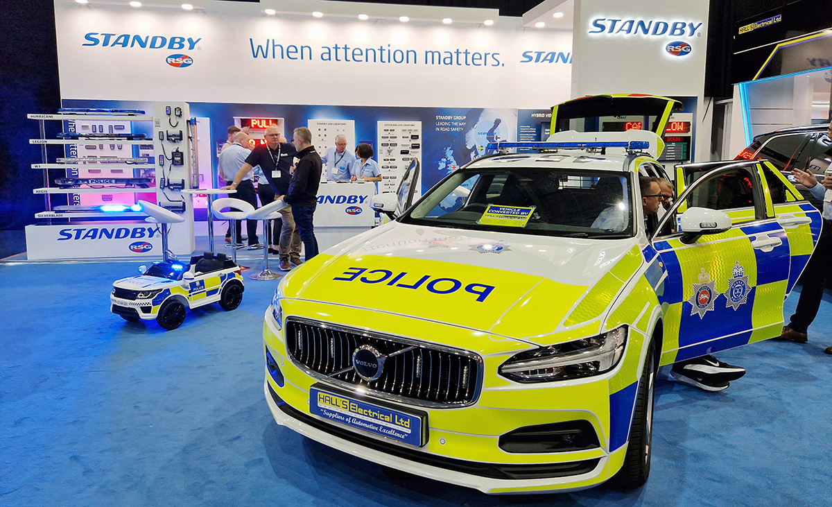 NAPFM Standby Stand Police Car next to children's ride-on RC car with blue flashing lights, various groups of people are stood around, person off screen talks to people in passenger and back of police car