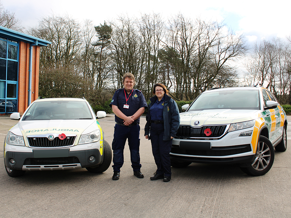 Two Community First Responders stand between two forward angle facing emergency response vehicles on an industrial park with foliage in the background. Both are facing the camera and smiling.