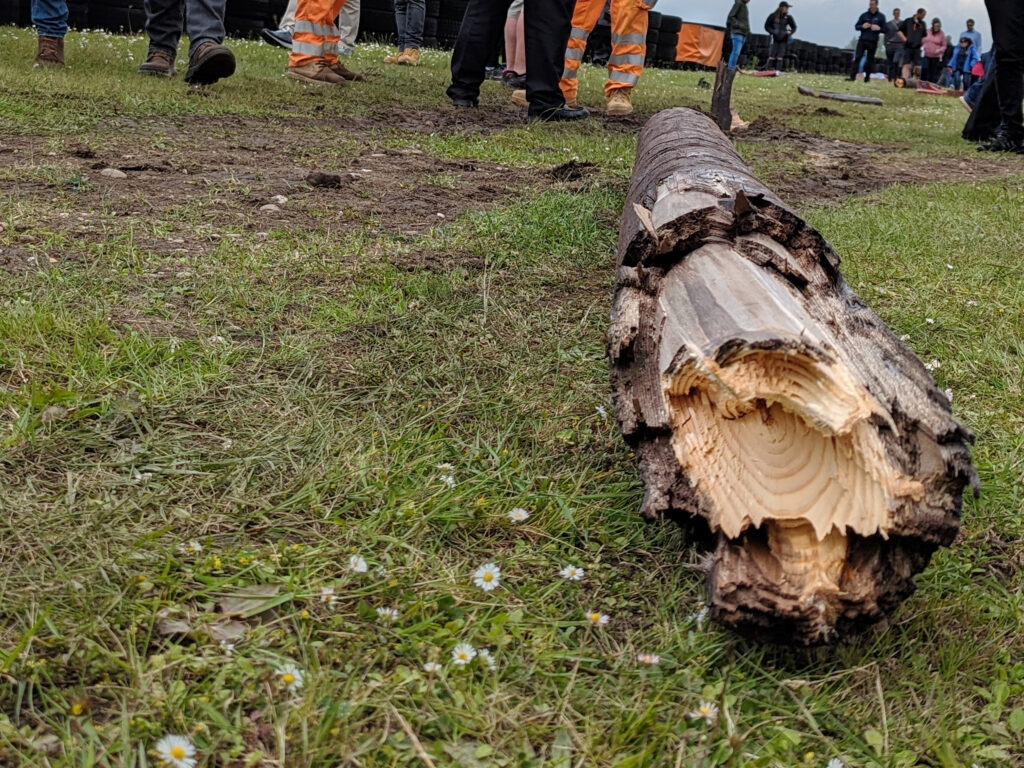 Damaged wooden pole lying flat on the grass, people's legs seen in the top of the image