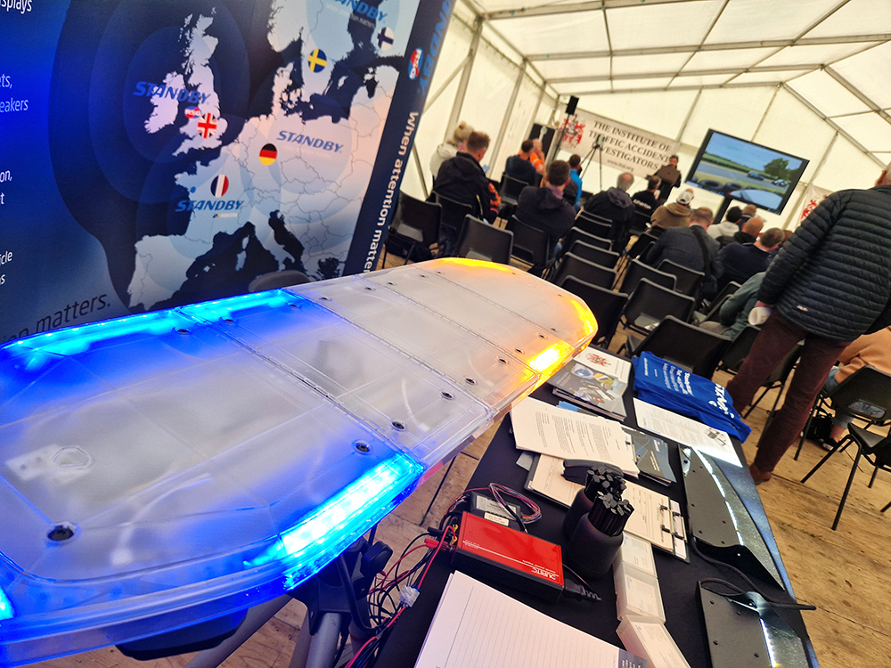 Angle shot of a small exhibition stand with a blue and amber lit lightbar. A crowd of people are seated in the background facing a screen and speaker. This takes place in a white marquee.