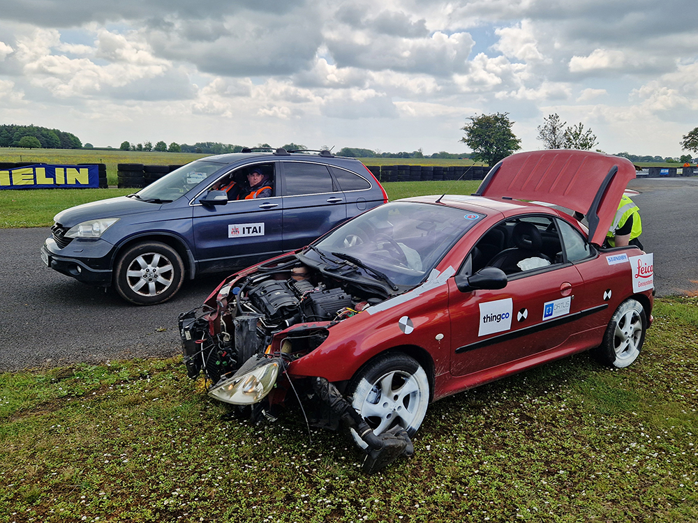 Heavilt damaged red coupe car with the bonnet missing and boot open on a racetrack. A blue vehicle is pulled up next to it with two men inside.