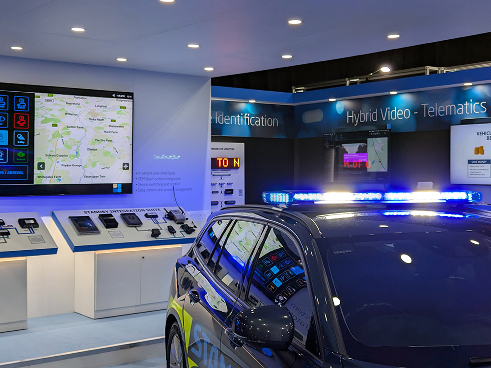 Close up shot of a lightbar lit up in blue and white on top of a dark coloured emergency response vehicle on an exhibition stand with a large screen in the background.