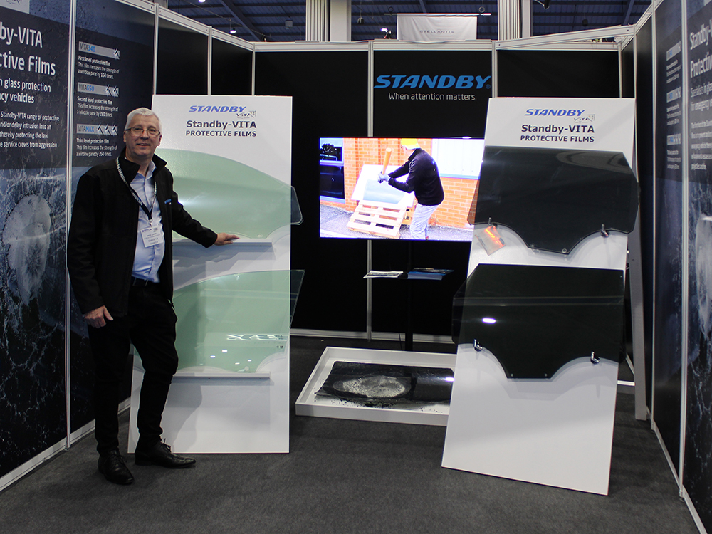 Simple black exhibition stand with a male Standby UK employee posed in front of stands with car windows. There is a screen in the middle of the stand, above a broken window in a tray. Stand branding reads 'Standby-Vita Protective Films'.