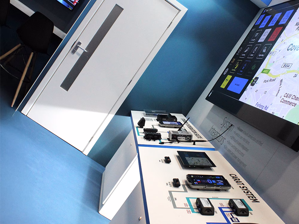 Cropped angle view of a large screen mounted on the wall of an exhibition stand with a door and counter with chair on the lefthand side.