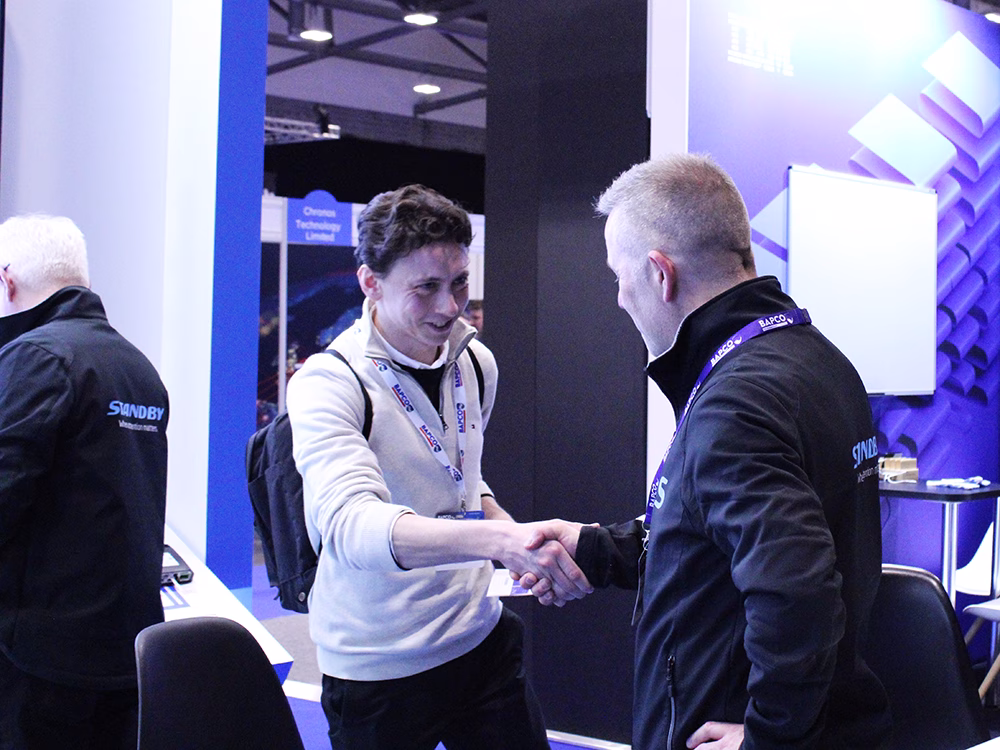 Standby employee shaking hands with a smiling stand visitor at an indoor exhibition, another standby employee has his back to the camera on the lefthand side.