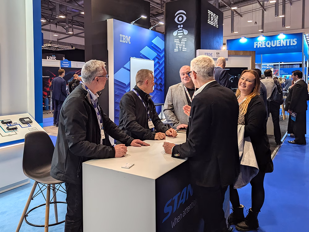 Group of people, some Standby employees and some stand visitors at a counter in an indoor exhibition. The female visitor is looking and smiling at the camera, all other people in conversation with eah other. The counter has Standby signage on.