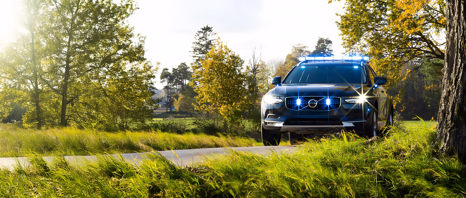 Standby Volvo with blue lightbar driving down a sunny road surrounded by plants and trees