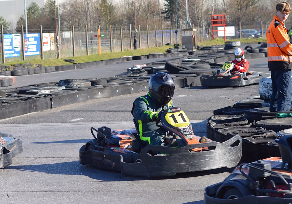 Racing karts on a track - number 11 and 10, the racers are wearing jumpsuits and helmets. A trak employee stands to the right of the tire wall in an orange hi-vis looking at the frontmost racer.
