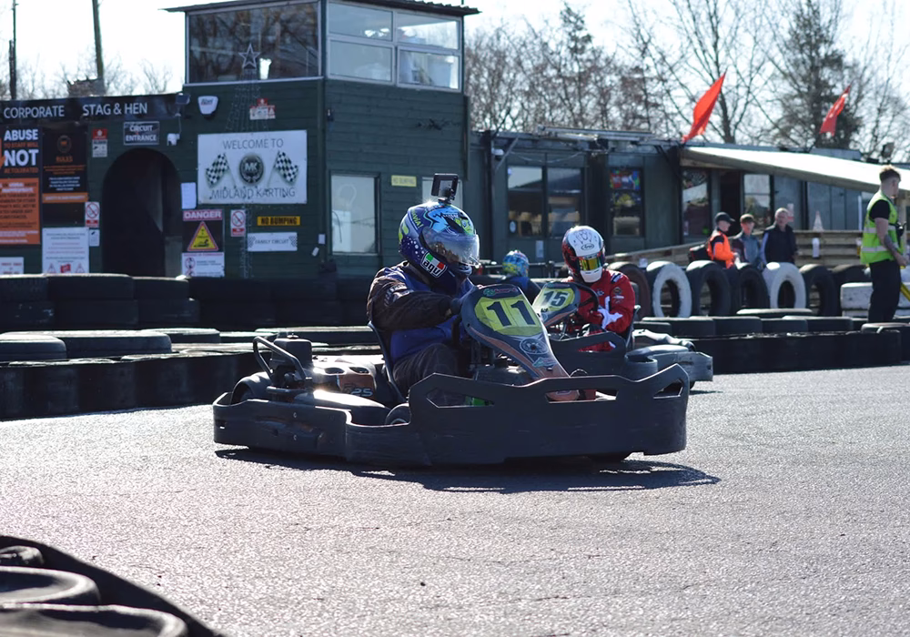Racing karts on a track - number 11 and 15 in front of a lookout building. The racers are wearing jumpsuits and helmets.