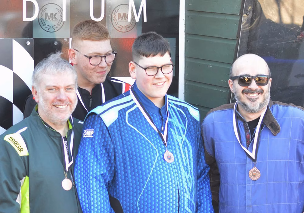 A team of 4 racers in jumpsuits wearing medals smiling, image cropped on their torso area.
