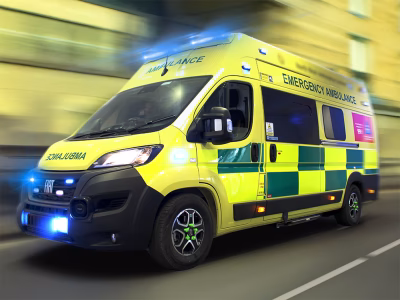 Side/front angle view of an abulance in motion, buildings in the background with motion blur. Blue flashing lights on the front of the vehicle.