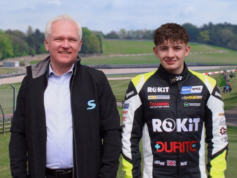 Standby UK's Sales & Marketing Manager Andrew Buxton stands next to BTCC Driver Max Hall, dressed in racing gear, in front of Donnington Park Racetrack.