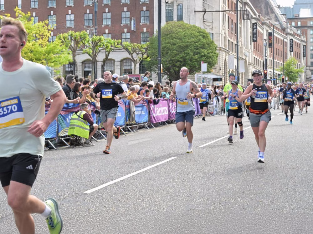 Runners running down a road cordoned off with barriers and lined with spectators in the city centre, flanked by trees and tall buildings.