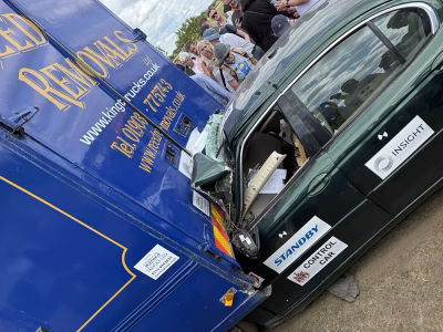 Close up angle view of a car collision at ITAI Crash Day 2025, the front section of the car is compacted underneath the back of a blue removals van. People crowd around to take photos. There are various company logos on the car including the Standby UK logo. Another decal reads 'control car' next to the ITAI crest.