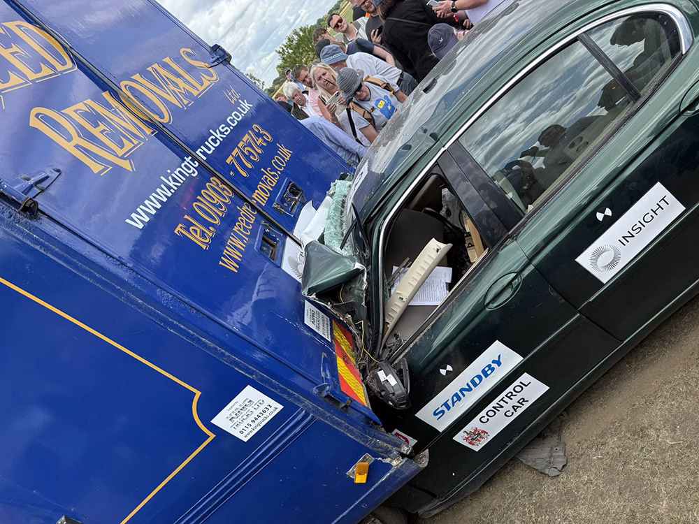 Close up angle view of a car collision at ITAI Crash Day 2025, the front section of the car is compacted underneath the back of a blue removals van. People crowd around to take photos. There are various company logos on the car including the Standby UK logo. Another decal reads 'control car' next to the ITAI crest.