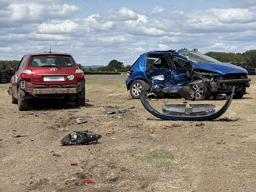 Aftermath of a car collision at ITAI Crash Day 2025. A red and blue car sit stationary on a patch of dried grass and mud. Both cars are badly damaged, the blue car has a crumpled side. There is debris lying across the floor.