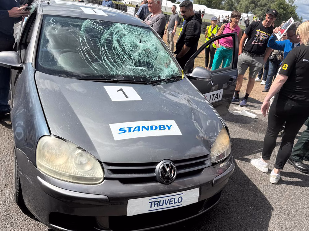 Dark silver car at at ITAI Crash Day 2025. It has a smashed windscreen and Standby logo on the bonnet. People crowd around to look, the passenger side door is open.