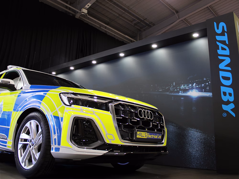 Low angle view of the front of a Police Audi Q7 against a black wall with vertical blue Standby logo on the Standby UK stand at NAPFM 2025.