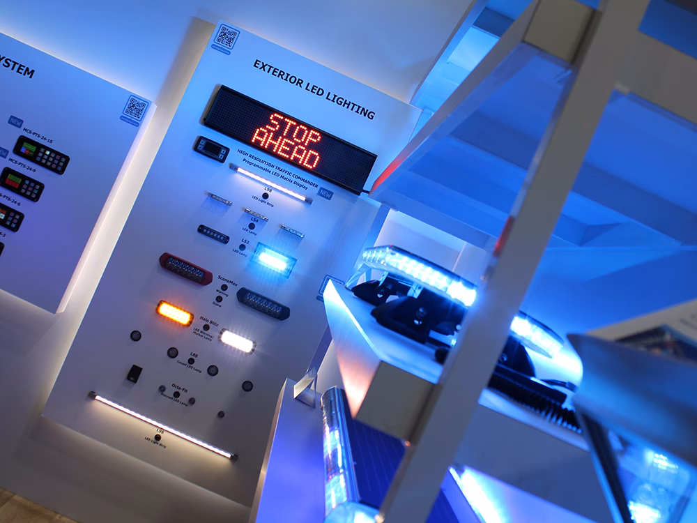 Angle view of Lightbars on shelves lit up blue and an Exterior LED Lighting panel with various lit and unlit products on the white Standby UK stand at NAPFM 2025. The blue lights are saturating the white surfaces.