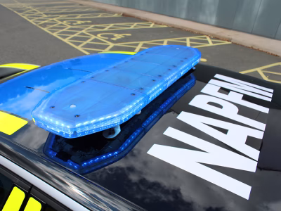 Blue Raiden lightbar on top of a black police vehicle lit up, taken at an angle with large white livery below reading 'NAPFM'. The car is parked outside in a car park in front of some disabled bays.
