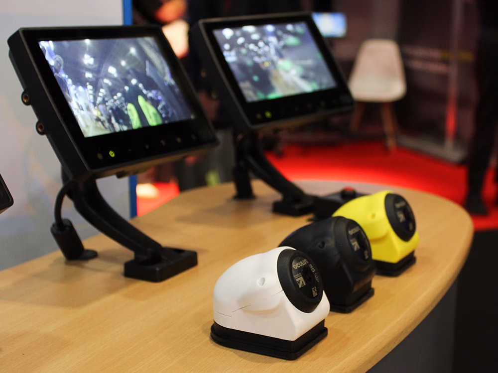 Angled shot of the Oculux cameras in white, black and yellow housing in front of some live feed monitors on the Oculux podium on the Standby UK stand the Emergency Services Show 2025.