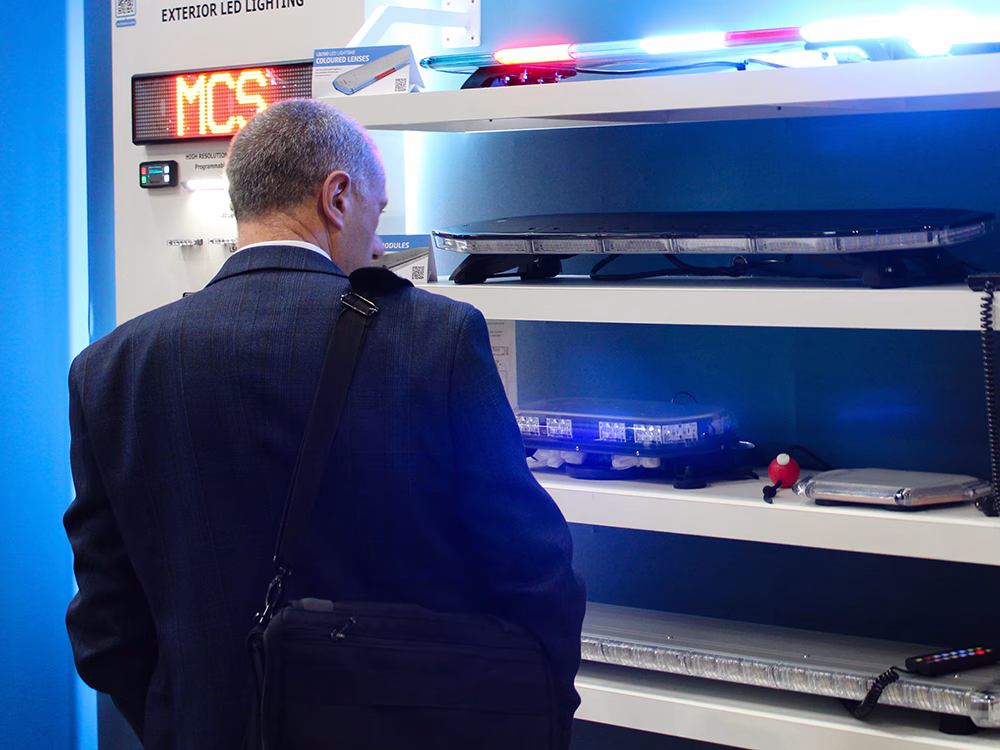 Man wearing suit stands in front of lightbar shelves on the Standby UK stand at the Emergency Services Show 2025 with his back to the camera