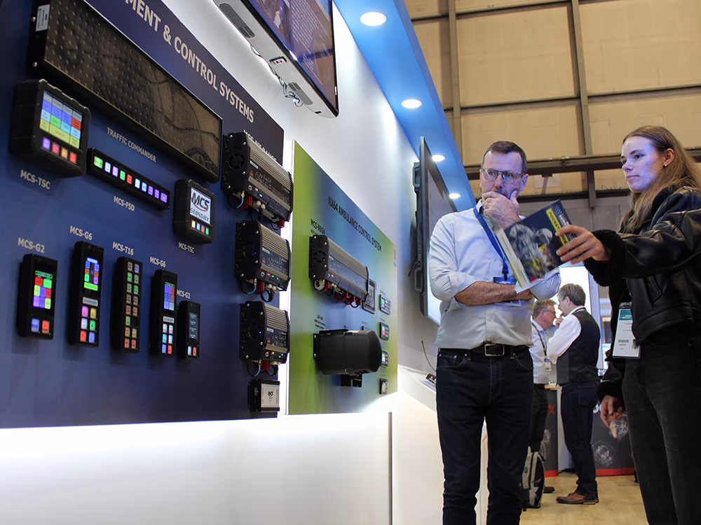 Stand visitors gestures at the Power Management & Control Systems board on the Standby UK stand at the Emergency Services Show 2025 whilst interacting with Managing Director Roy McClarty.