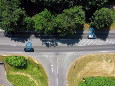 Aerial view of a road junction in the UK between trees with blue location pins over some of the vehicles on the road.